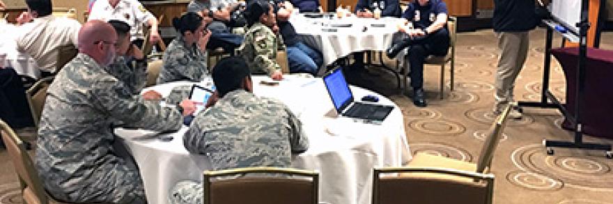 A man writes on a easel at the front of a room filled with onlookers, some of whom are in military dress.