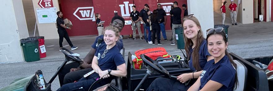Four Intra-Collegiate Emergency Medical Service (IC-EMS) students smiling at the camera from electric carts while on duty at the Indiana University football stadium. 