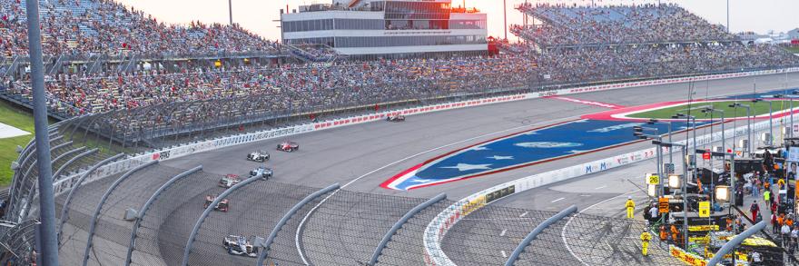 Race cars driving on the Iowa Speedway’s seven lane, oval racetrack in Newton, Iowa with thousands of spectators sitting in the surrounding stadium.
