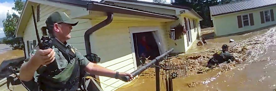 A first responder in tactical gear and holding a radio uses a pole and rope to assist a person through severe floodwaters outside a damaged home; Debris and muddy water surround the homes 