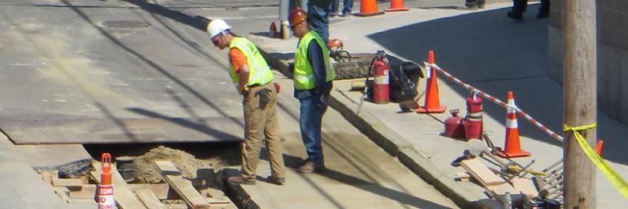 Workers in safety vests and hard hats observe a hole in the street.