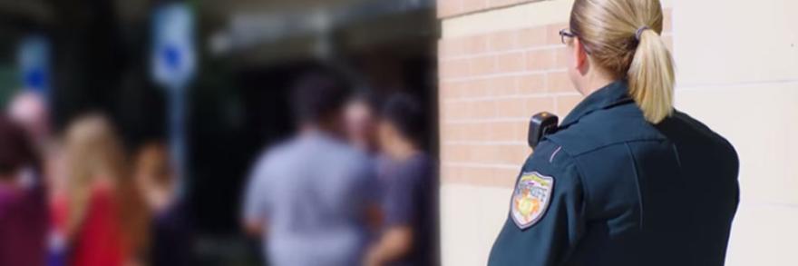 A female Sheriff’s deputy stands next to a brick wall and watches over a group of students.