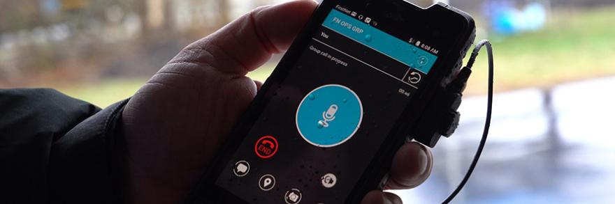 Hand holds FirstNet smartphone device with push-to-talk app displayed.