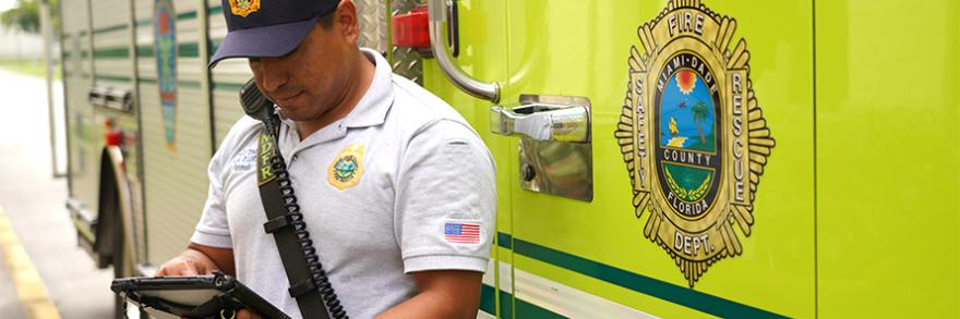Fire fighter standing in front of a fire apparatus wearing a radio while typing on a mobile device.