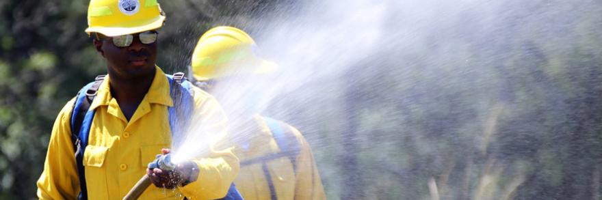 A first responder in a hard hat holds a hose spraying water; a second first responder in a hard hat stands behind.