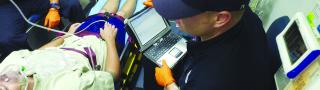 An EMS technician enters patient information into a laptop while riding in the back of an ambulance with a person in a stretcher