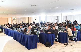 People sit at tables in a crowded room, working on their laptops and listening to a presenter.