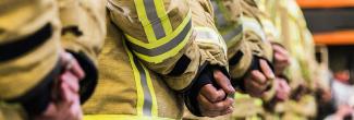 Firefighters stand in a line with their hands clasped behind their backs.