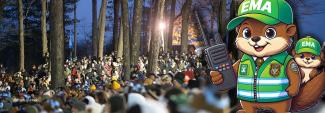 A group of people gathered in a wooded area attending the annual Groundhog Day ceremony. A cartoon image of groundhogs dressed as emergency management officials with one holding a mobile device.