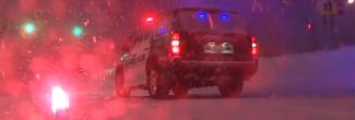 A police SUV with lights on and lit flare sit in a snowstorm at night.