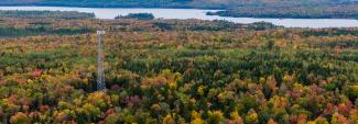 Aerial view of cell tower in a rural, forested area in autumn; a body of water is visible behind the tower