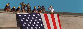 A group of firefighters in helmets and military personnel in fatigues, some saluting, standing on the roof of the Pentagon in Arlington, Virginia, unfurling a large American flag over the scarred stone.