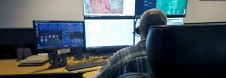 911 call-taker wearing a headset sitting at a desk in a communications center; multiple computer monitors showing maps, data, and emergency calls