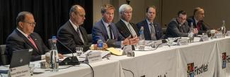 Seven of the FirstNet Authority's Board members sit at tables during a meeting in front of microphones