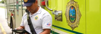 Fire fighter standing in front of a fire apparatus wearing a radio while typing on a mobile device.