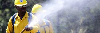 A first responder in a hard hat holds a hose spraying water; a second first responder in a hard hat stands behind.