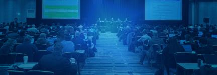 A large conference room filled with rows of people seated at long tables, facing toward five presenters sitting at a dais and two jumbo screens in front of them at a 3GPP meeting