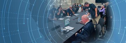 Attendees gather around a table at the FirstNet Users Summit; abstract pattern moves outward from attendees like rings on a tree