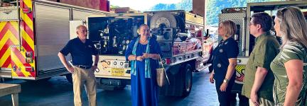 The FirstNet Authority’s Alexandra Fernández Navarro and Michael Varney and others talk with an official from the U.S. Pacific territories, standing at the rear of three public safety vehicles in an open apparatus bay.