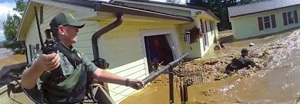A first responder in tactical gear and holding a radio uses a pole and rope to assist a person through severe floodwaters outside a damaged home; Debris and muddy water surround the homes 